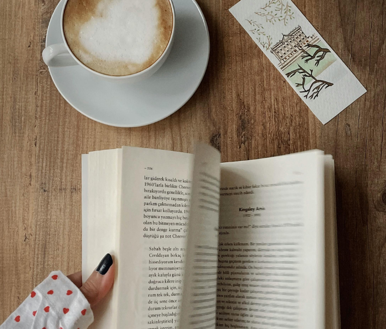 A woman's hands, turning the pages of a book on a wood table with a cup of cappuccino nearby