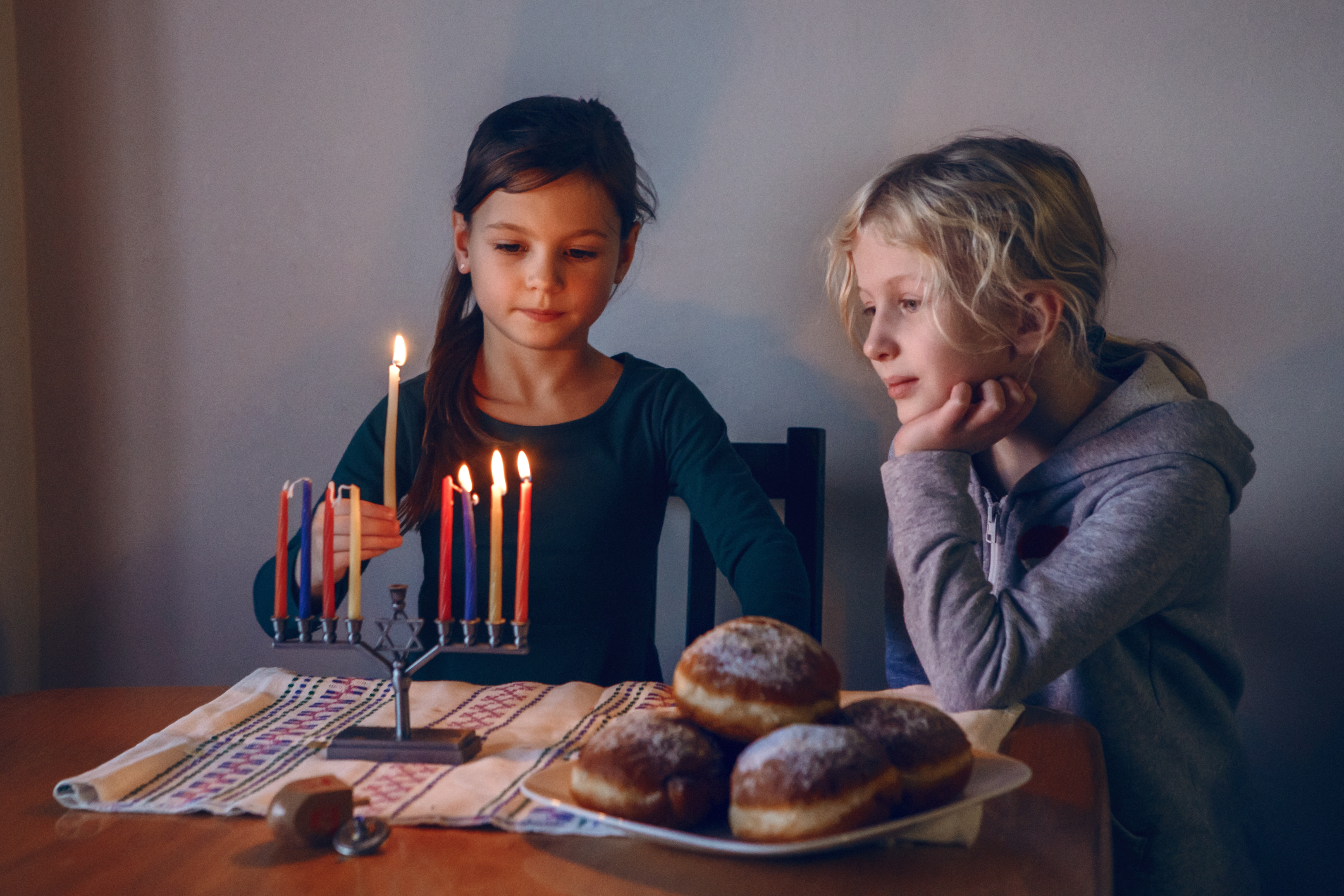 two young girls lighting a menorah on a table with donuts and a dreidel