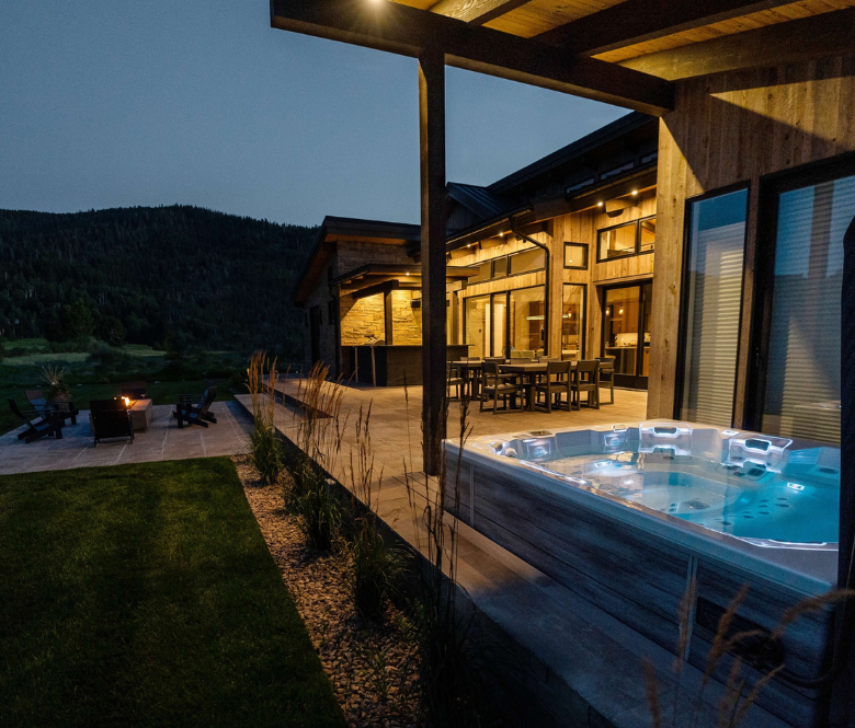 A backyard with a lit up spa and firepit surrounded by grass and trees.