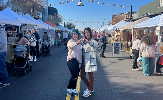 Women standing in the middle of a vendor fair street holding their hands together in a heart shape.