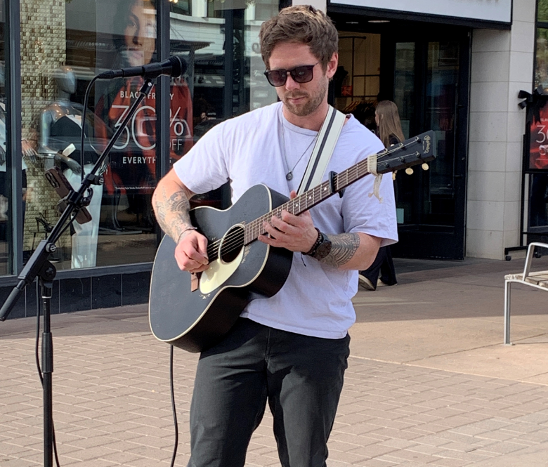 Musician holding a guitar in front of a microphone with a store in the background.