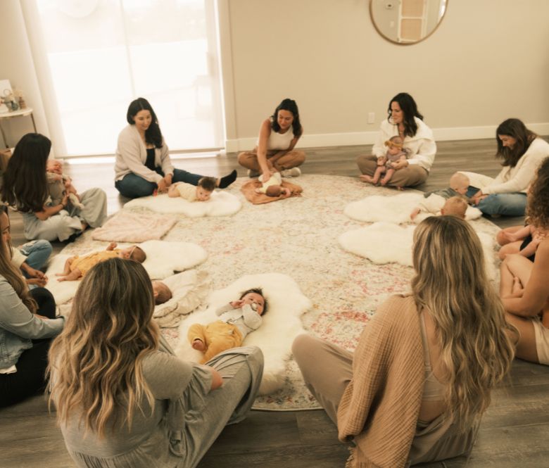 People sitting in a circle with babies laying on rugs in front of them.