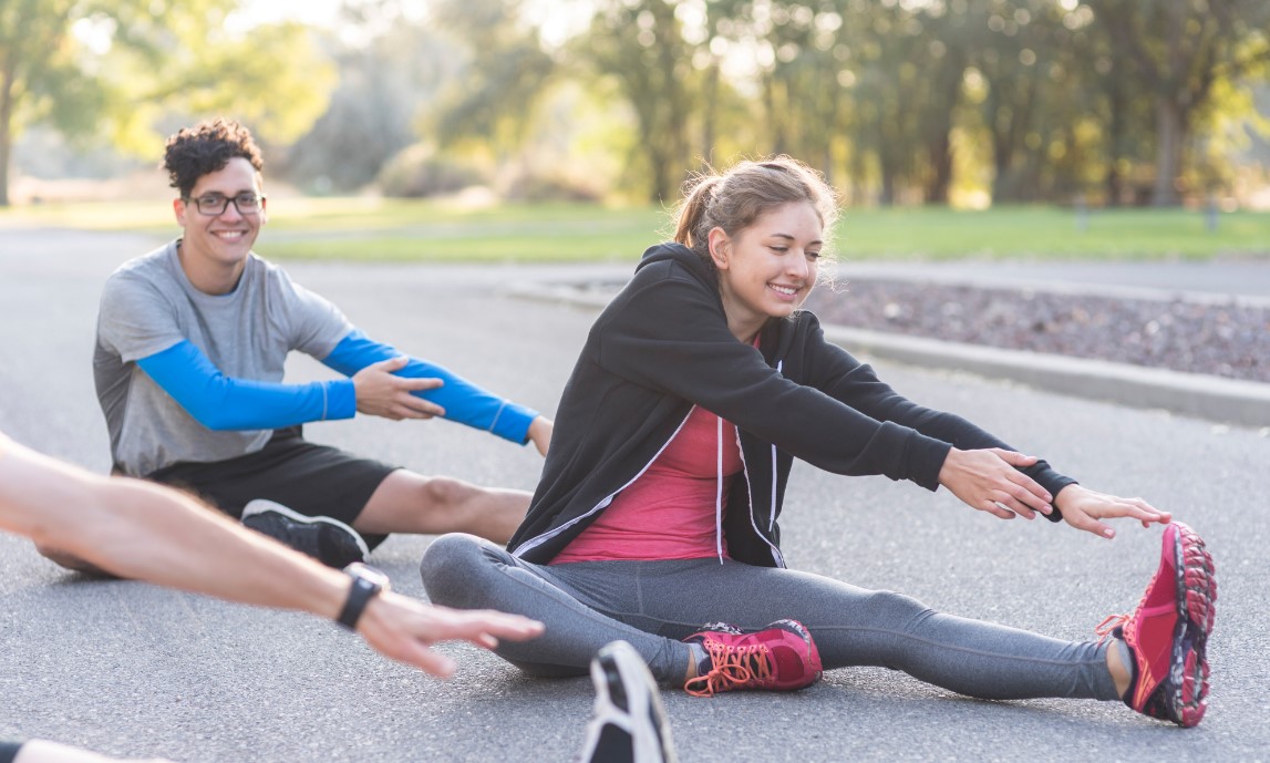 Two people in workout clothing sitting on the ground stretching their legs.