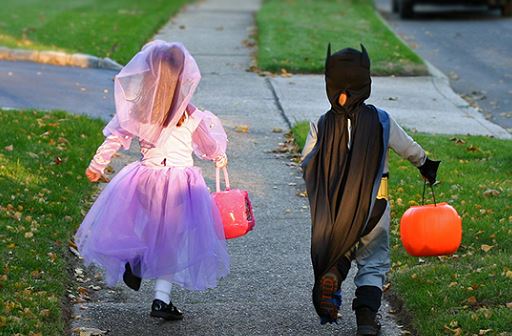 image from behind of 2 children walking down the sidewalk with their candy bags for Halloween trick or treating. Female child dressed in pink and purple princess dress with bright pink candy bag and male child in grey and blue batman costume with black cape and orange jack-o-lanter pumpkin for candy bag.