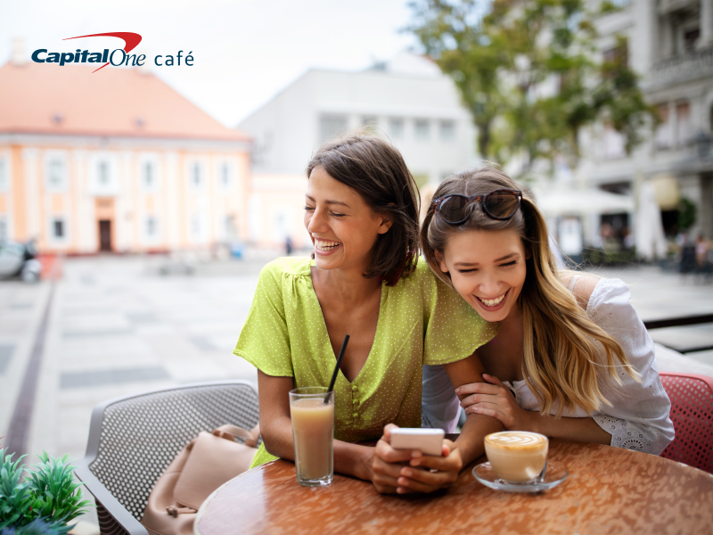 TWO GIRLS SITTING AT A TABLE LOOKING AT A PHONE LAUGHING OVER COFFEE