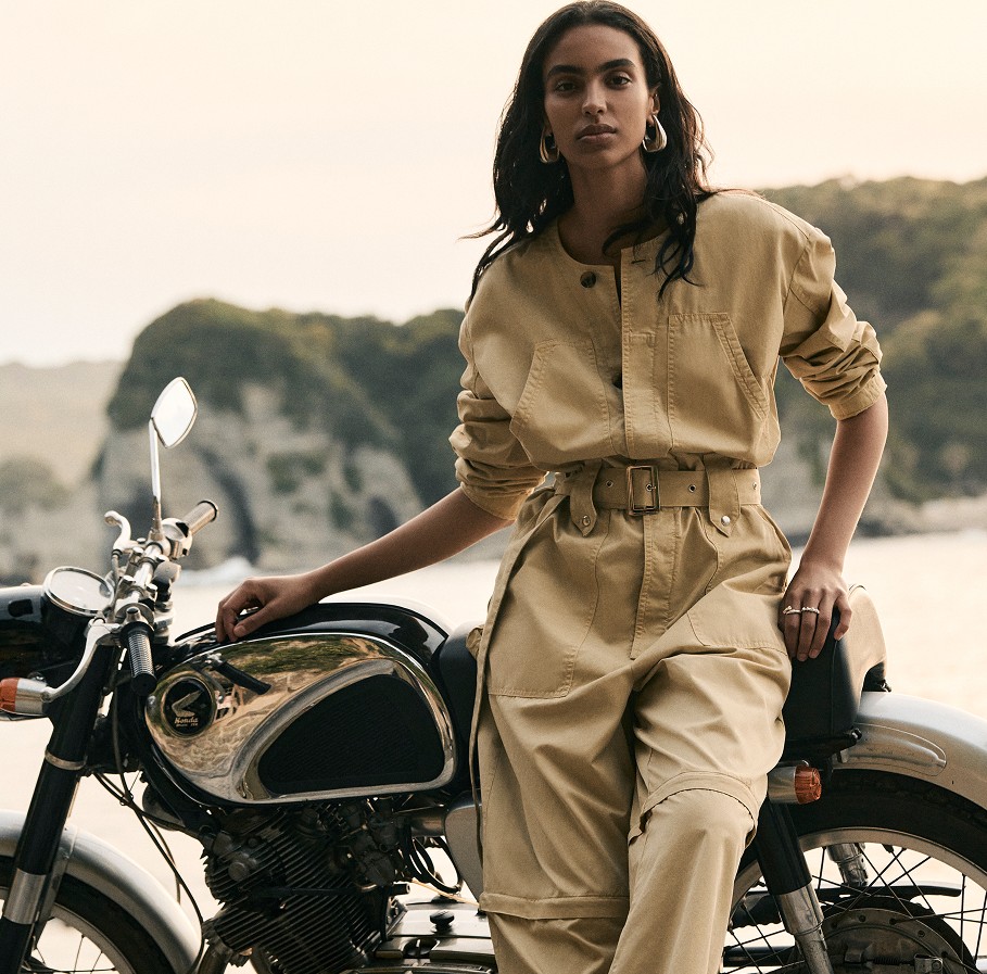 female model in a tan jumpsuit posing on a motorcycle