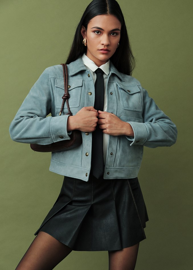 female model wearing a blue suede jacket, denim pleated skirt, white button-down with black tie
