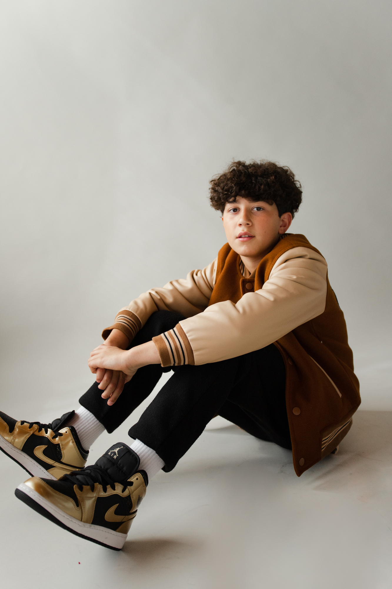 male model wearing varsity jacket black jeans and gold and black nike sneakers sitting on the floor posing