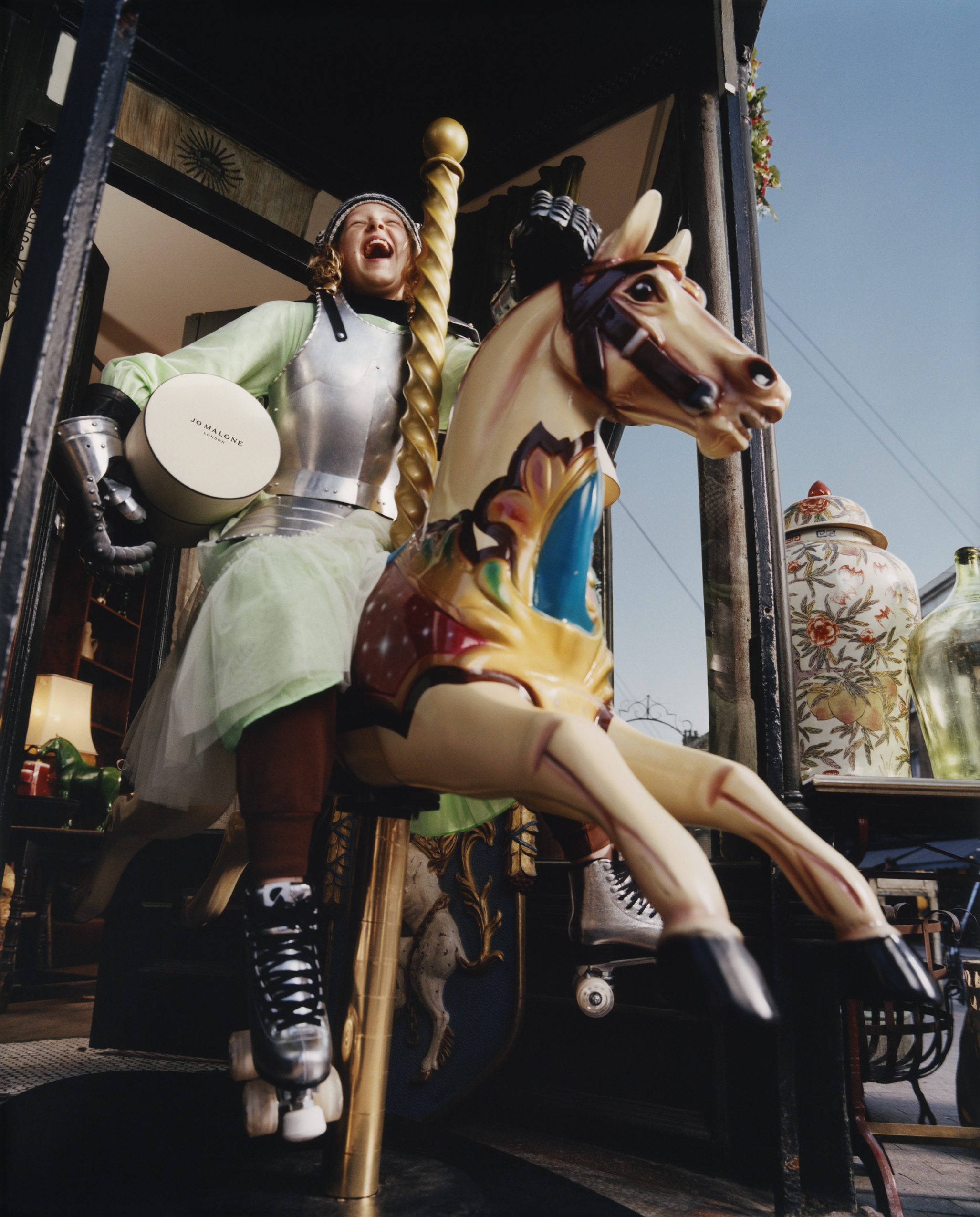 young girl with a green dress with silver rollerskates on holding a jo malone box riding a merry-go-round