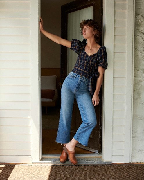 Girl standing in the doorway with blue jeans on with a plaid top with brown shoes. 