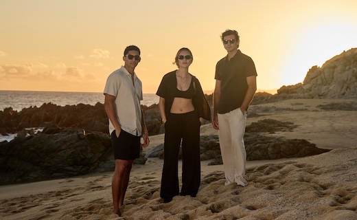 2 males and 1 female model wearing sunglasses standing on a beach in breezy light clothing