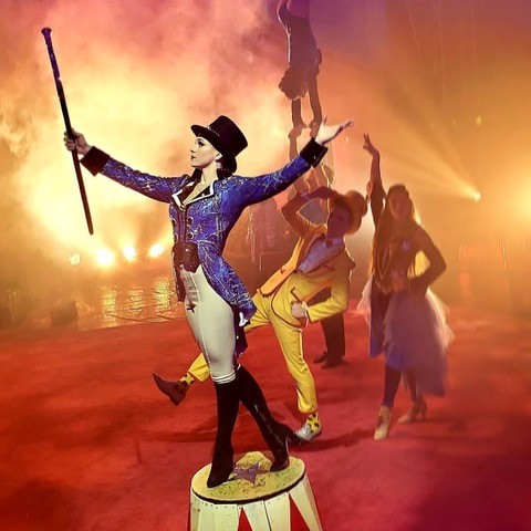 Ring master standing in front of a circus with man in suit, lady in skirt, man carrying lady upside down by the hands 