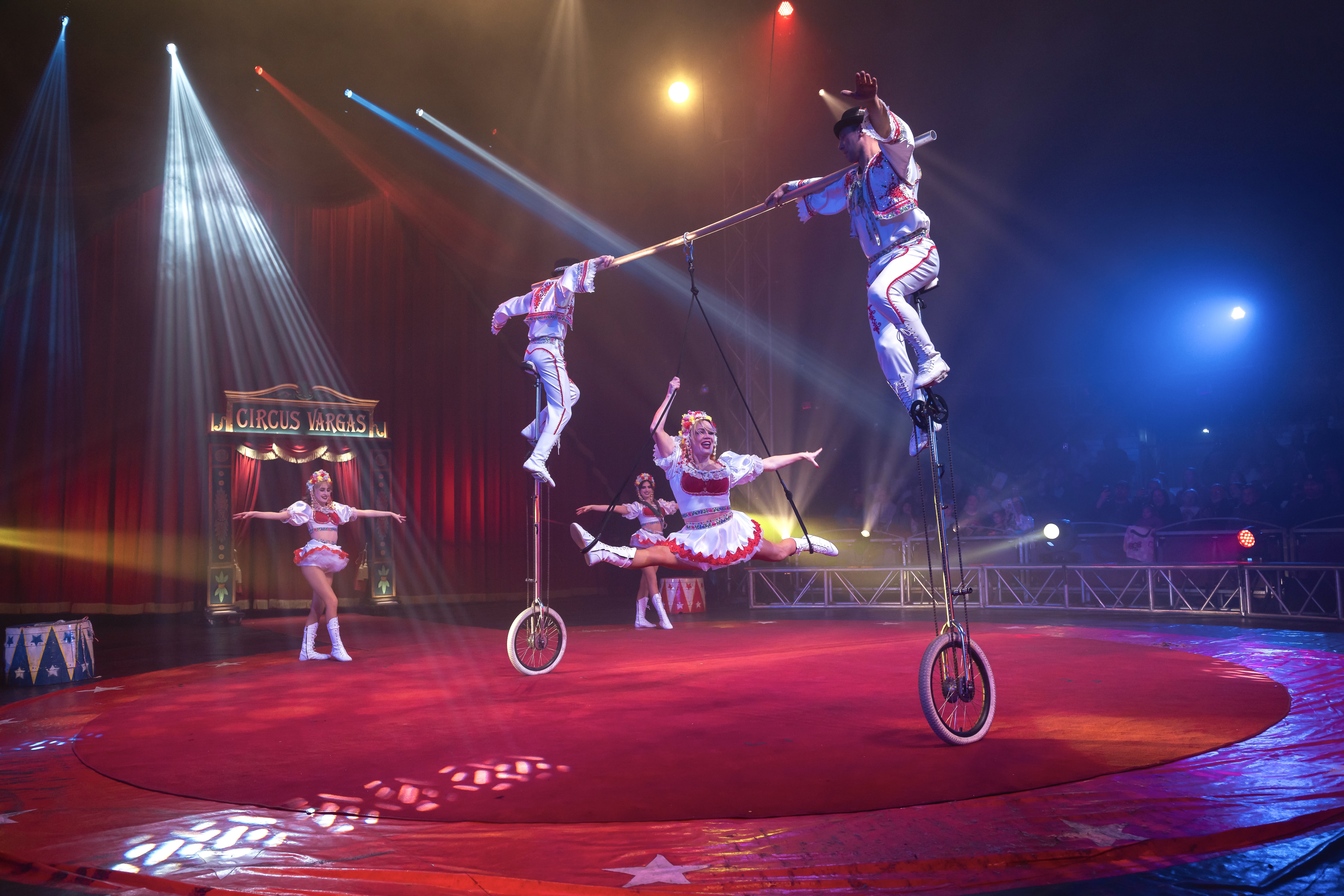 two men on a unicycle holding onto a woman. There are two other women watching in colorful costumes.