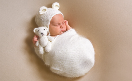 Baby sleeping and wrapped in white blanket and white cap with ears and holding a white teddy bear. 