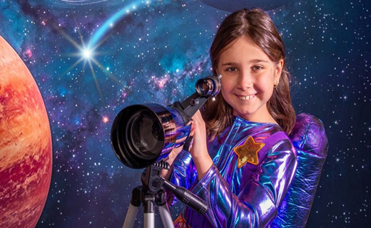 Girl dressed in a purple and blue spacesuit holding a telescope viewing the stars and planets in the distance. 
