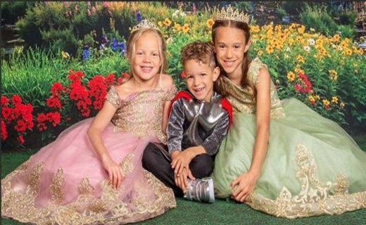 Two girl dressed as princess, one in a pink gown and one in a green gown, both with crowns on their head with a little prince in the middle of them all sitting in front of a lush green, floral garden with flowers in red, blue, orange, white and pink.