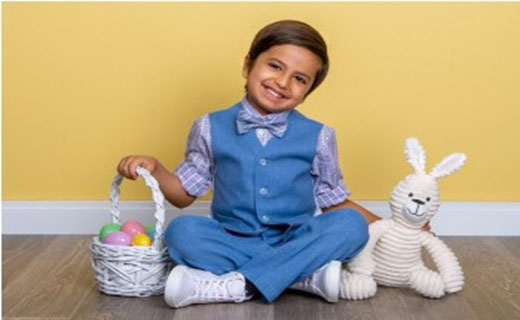Boy dressed in blue vest and blue pants with pinstriped bowtie and shirt holding Easter basket filled with Easter eggs in one hand and stuffed Bunny in one hand.