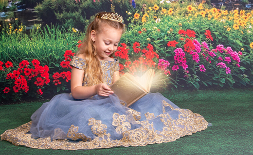 Little girl in a blue lace princess dress with tiara in her hair sitting and reading a magical book on the grass with a beautiful floral background filled with lush greenery, trees and red, pink, and orange flowers. 