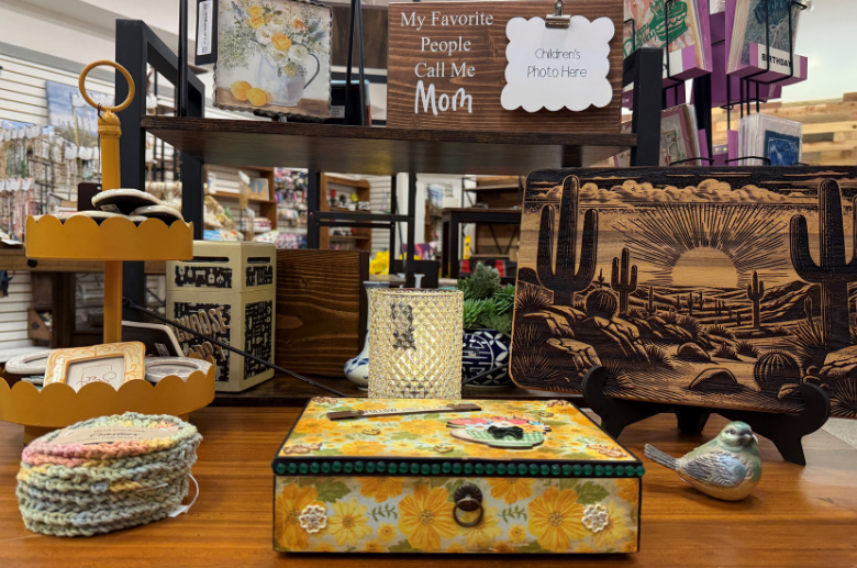 A table with various gift items including a wooden desert landscape picture, ceramic bird, crystal candle, curio box, crocheted coasters and more. 