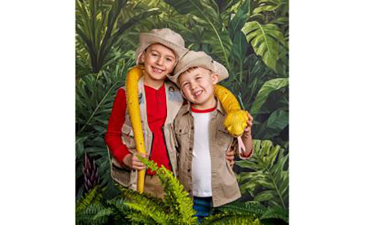Two young boys with safari hats and jacket with yellow snake around them and green foliage background