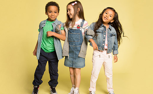 Boy wearing green t-shrit and black pants, interlocking arms with girls in denim dress and girl in denim jacket smiling and looking at the camera. Yellow background. 