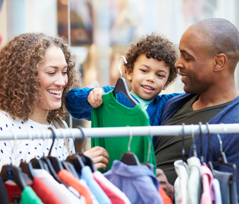 A family enjoys shopping together, smiling as their young child picks out a green shirt from a clothing rack.