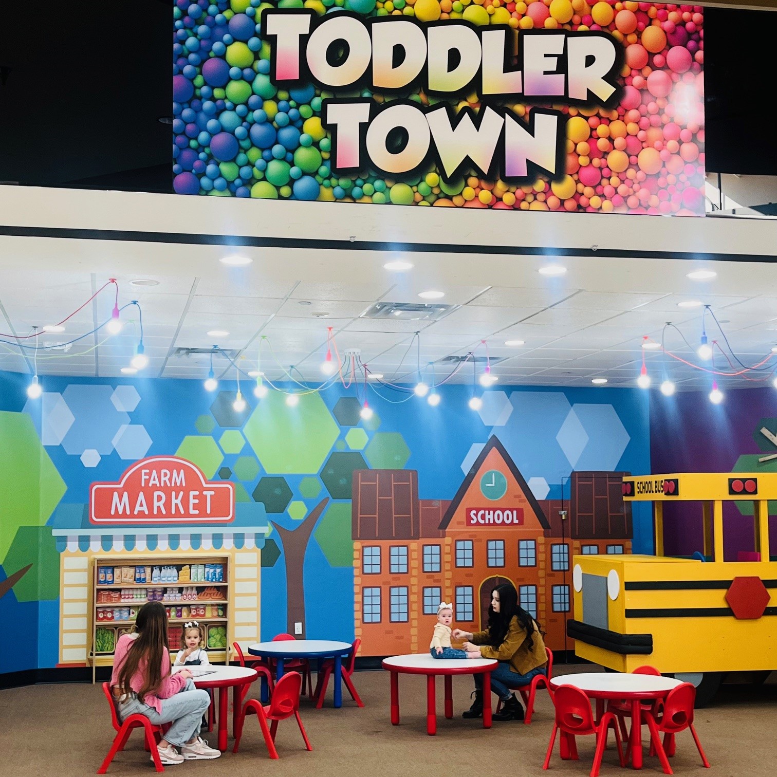 Two moms sitting with their kids in a kids space called "Toddler Town" that looks like a mini town. 