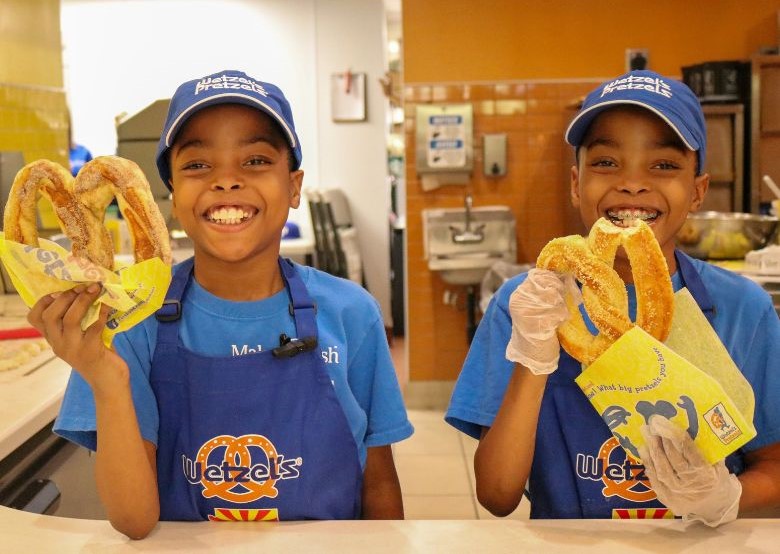 Two smiling children wearing Wetzel's Pretzels caps and blue aprons, each holding a large twisted pretzel, standing behind a counter in a Wetzel's Pretzels kitchen. 
