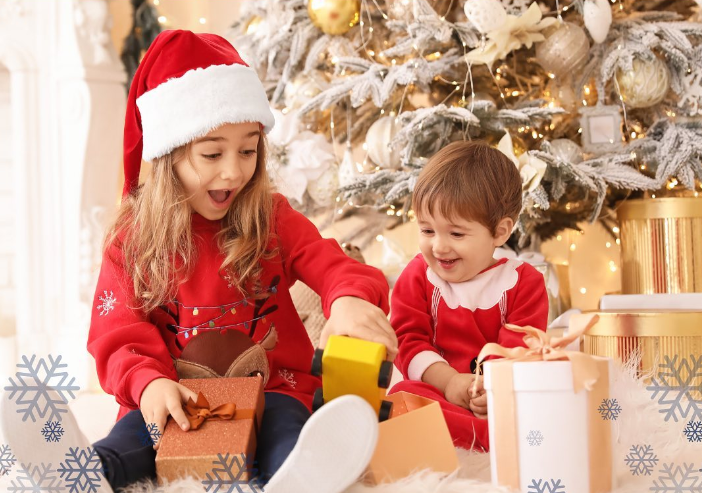 Two kids opening presents by a Christmas tree. Young girl wearing a Santa hat.