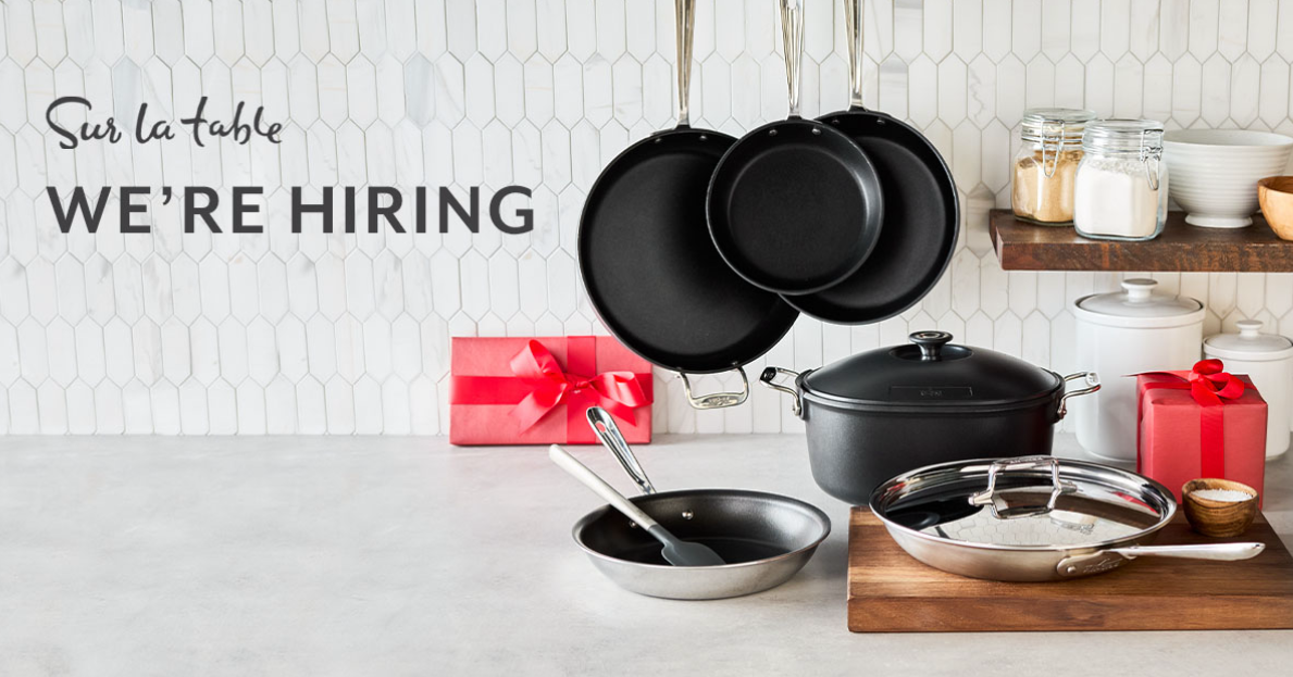 Kitchen items such as pots, pans, cutting board, etc. and two red presents wrapped, sitting on a countertop. Copy reads "Sur La Table. We're Hiring".