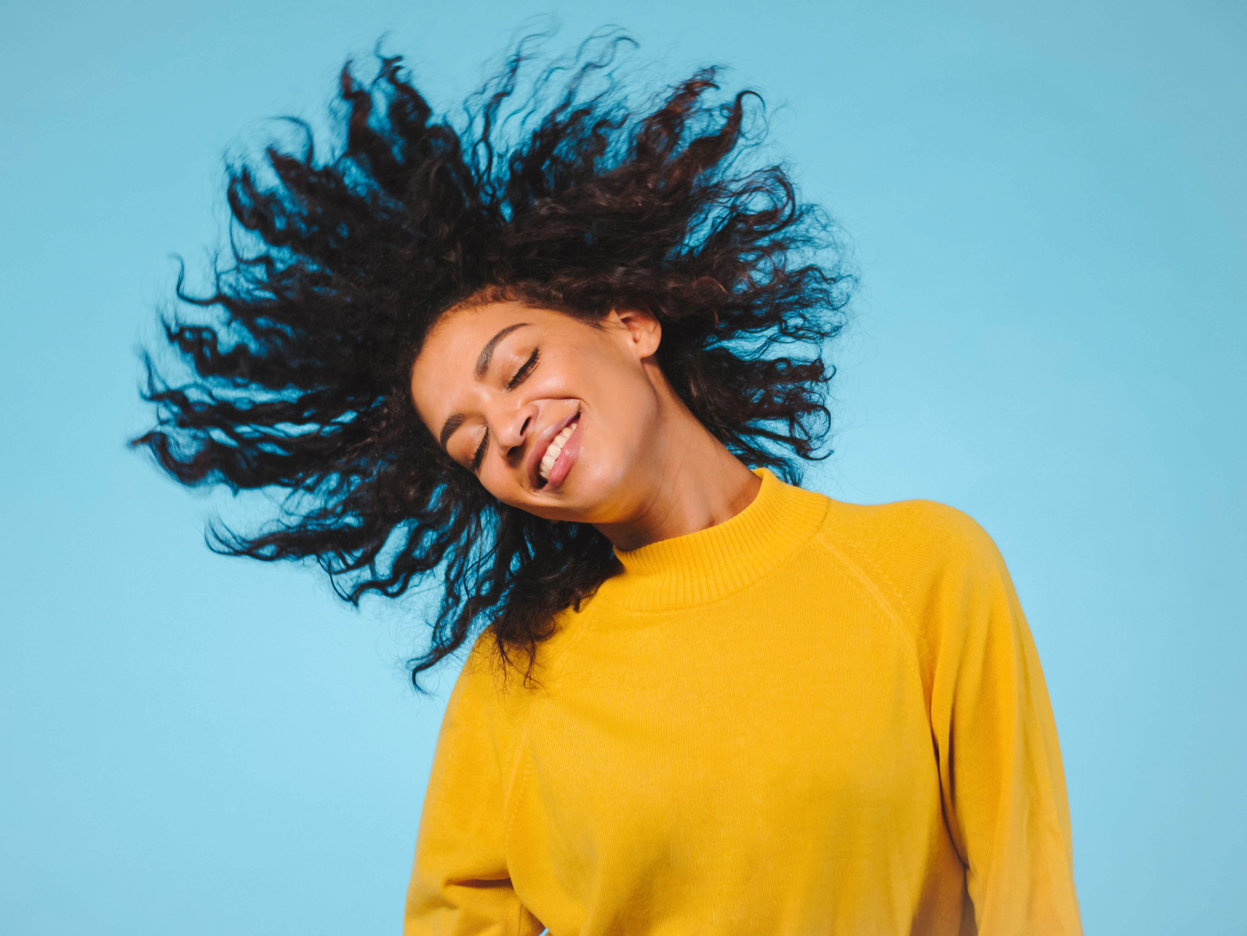 Person wearing a bright yellow sweater tosses curly hair against a light blue background.