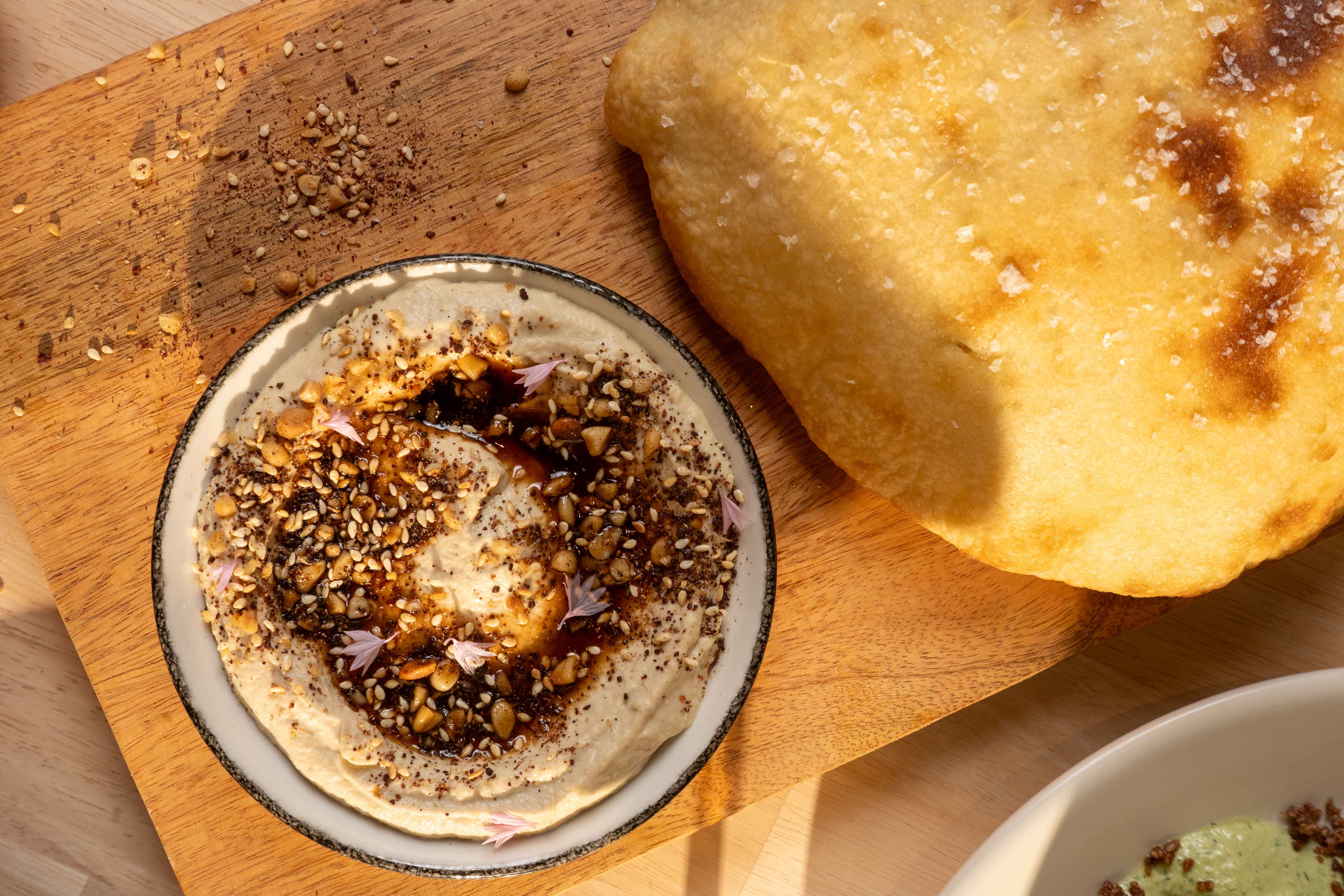 Bowl of hummus topped with seeds, spices, and olive oil, served alongside warm pita bread on a wooden board.