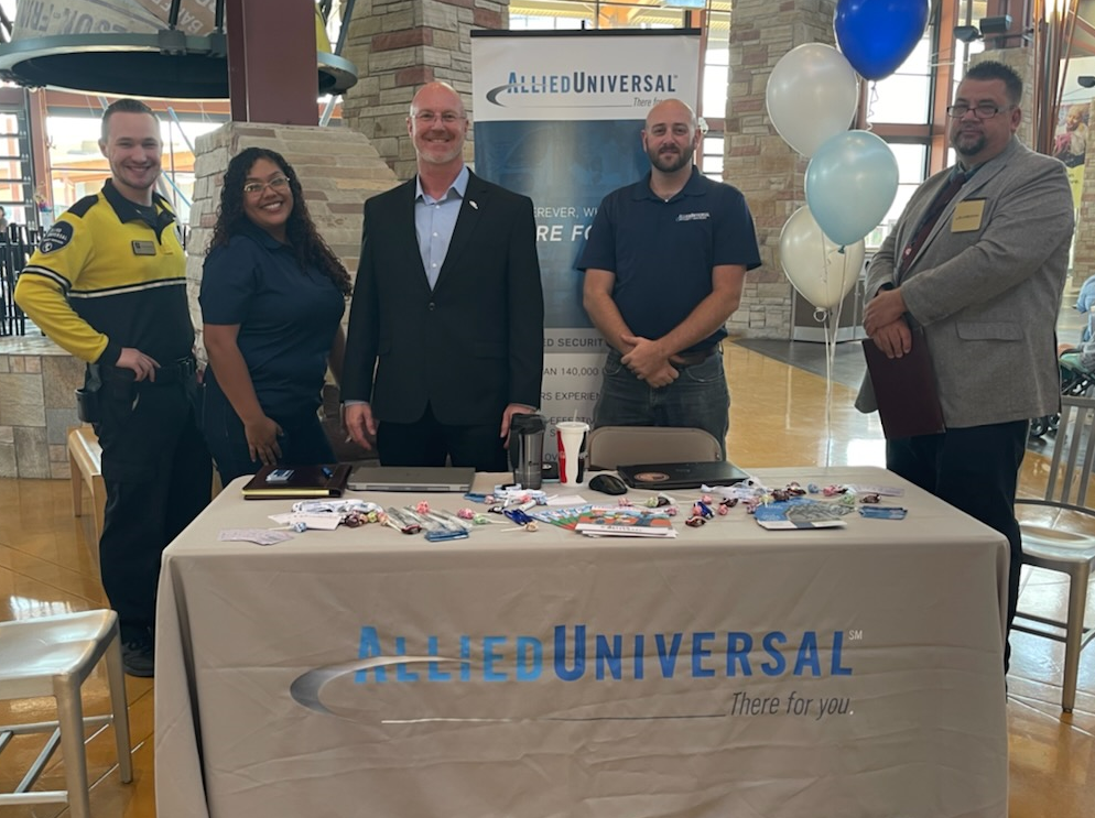 5 people standing in front of a promotional table. Text reads ' Allied Universal. There for you'