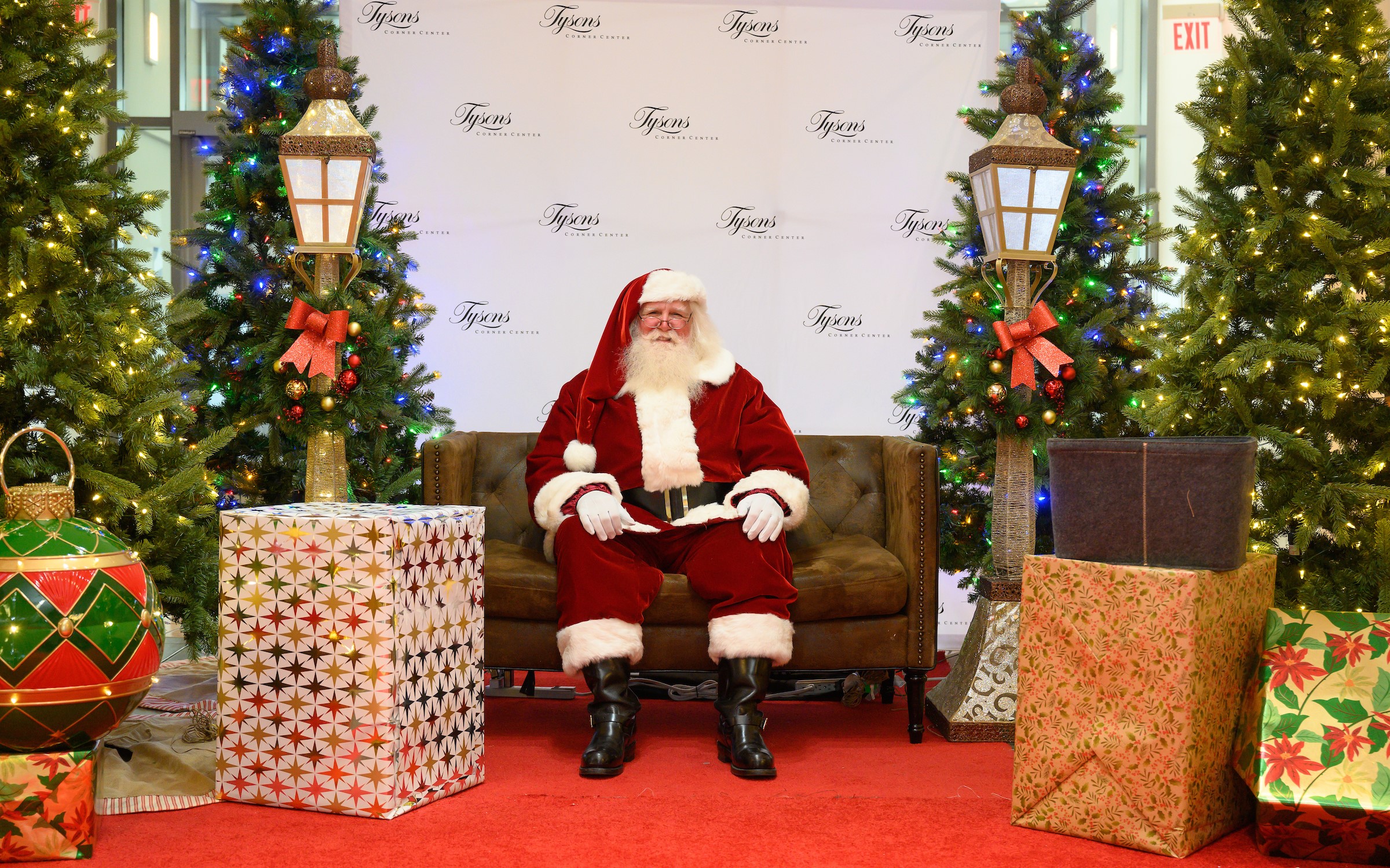 Santa seated in large chair surrounded by wrapped gifts and lit pine trees