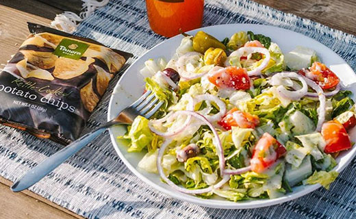 Salad on plate, bag of chips, and beverage on place mat in the sun.