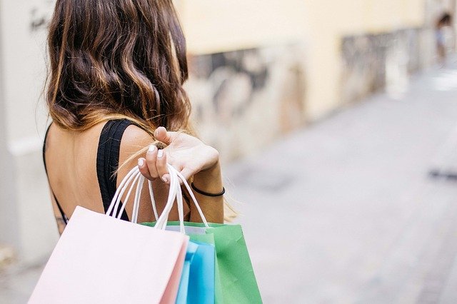 A woman holding a pink, blue, and green bag in her hands over her shoulder while walking down a street.