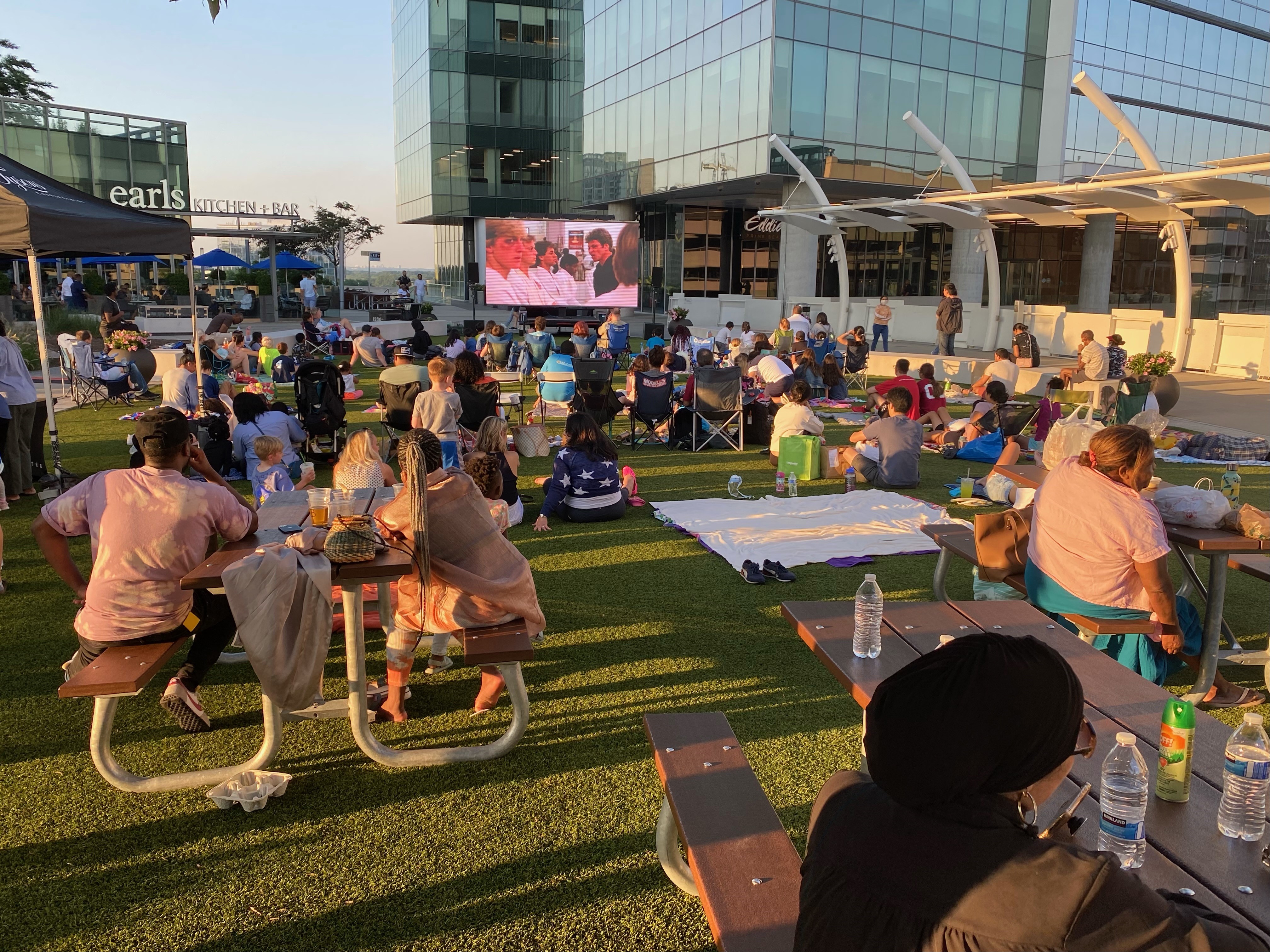 groups of families and friends spread out on the lawn watching a movie on a large screen. 