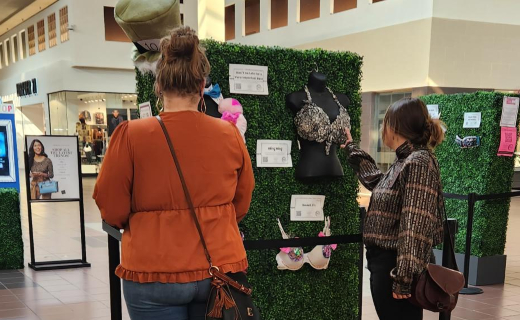 two women looking at decorated bras on display inside of a mall.