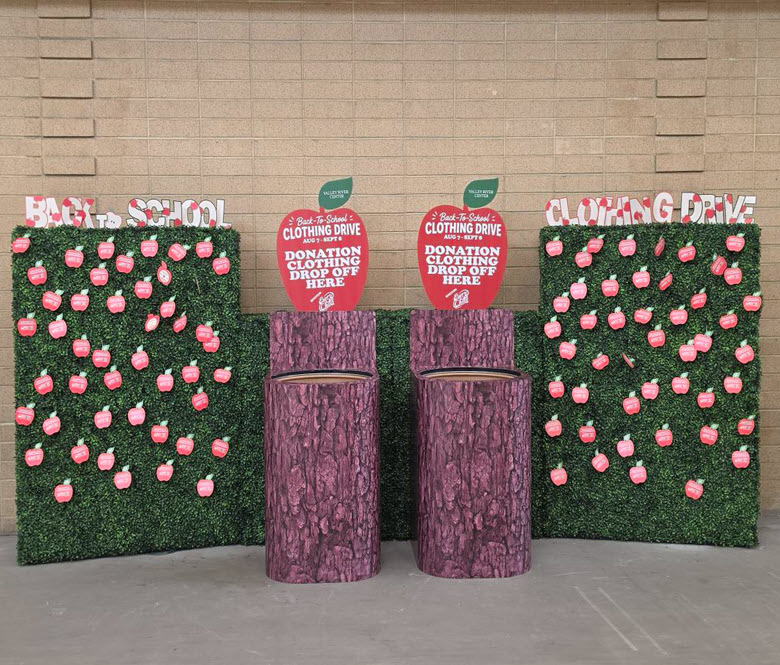A green hedge display decorated with red apple-shaped donation tags stands inside Valley River Center. Two round donation bins are positioned beside the hedge for clothing contributions.
