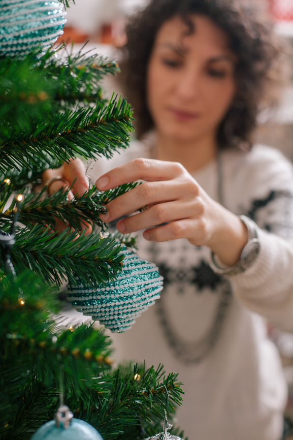 A woman putting a sparkling teal and white ornament on the branch of a green tree.