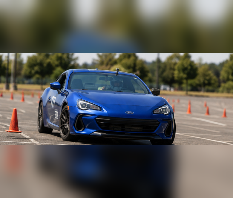 A blue sports car navigates an autocross course marked with orange traffic cones in a large outdoor parking lot.