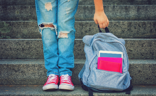 The lower half of a person wearing a pair of torn up jeans, a pair of red tennis shoes and their hand grabbing a blue backpack.