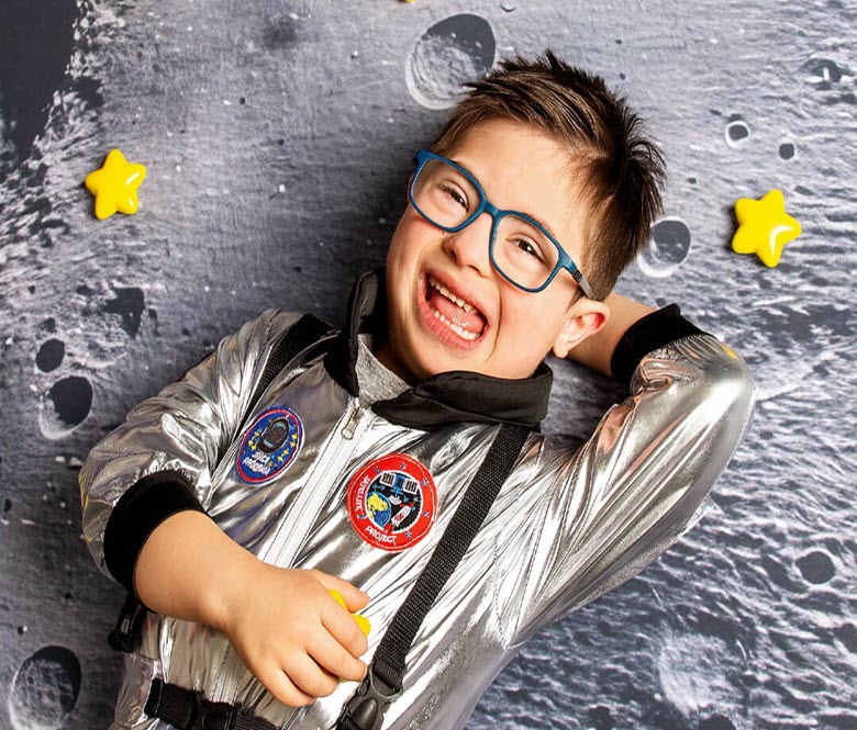 A smiling child wearing glasses and a silver astronaut costume poses in front of a space-themed backdrop with stars.