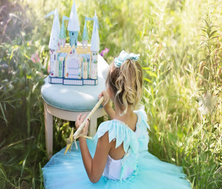 A little girl in a princess dress, holding a wand and playing outside with a toy castle.