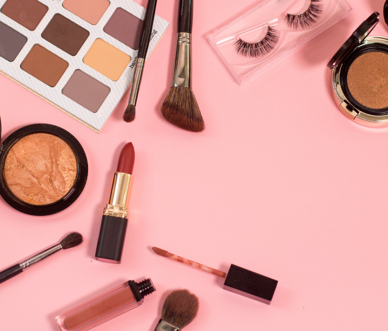 lipstick, lip gloss, eye shadow and some makeup brushes displayed on top of a pink background.