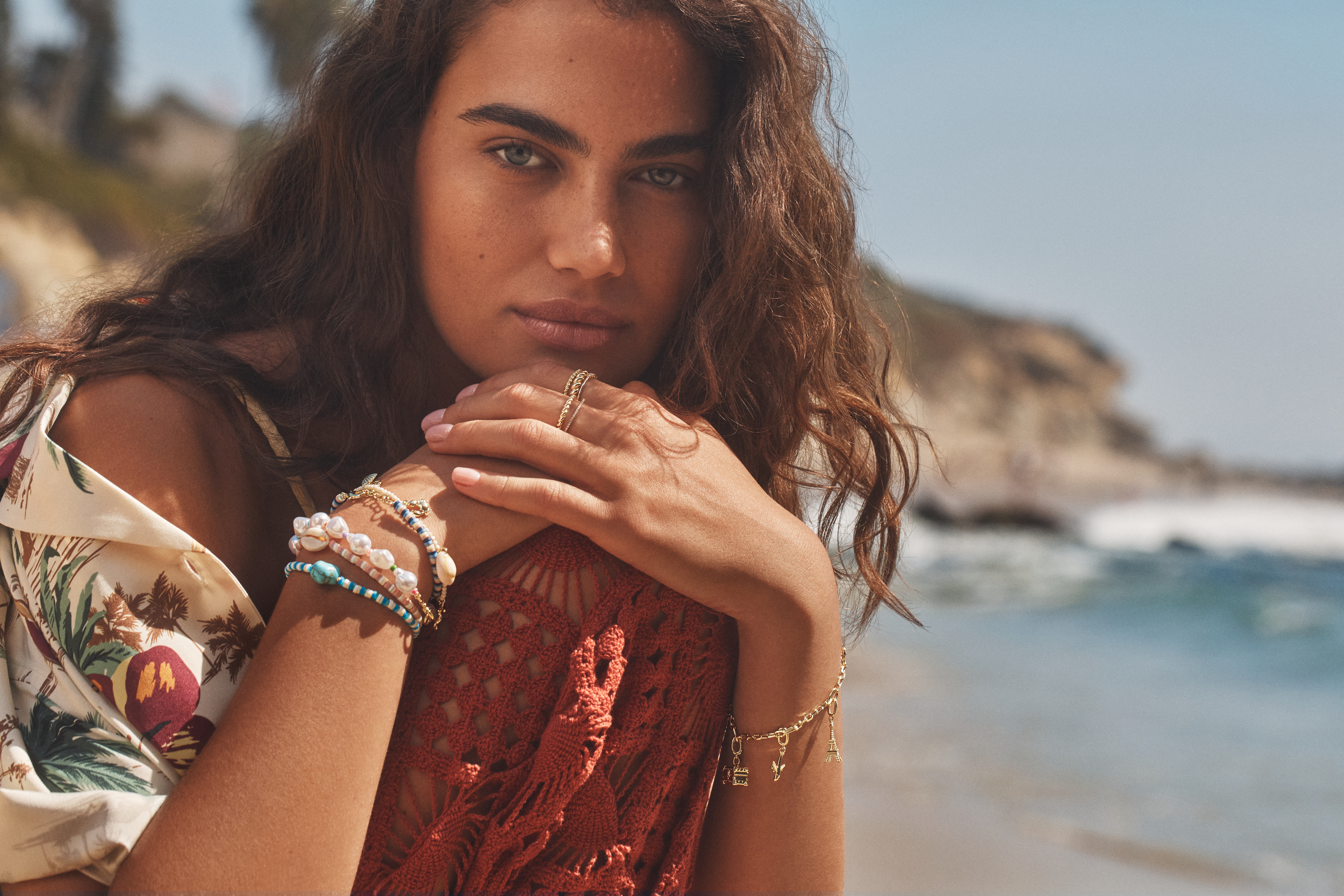 A woman wearing a floral blouse and pearl bracelets with the beach in the background
