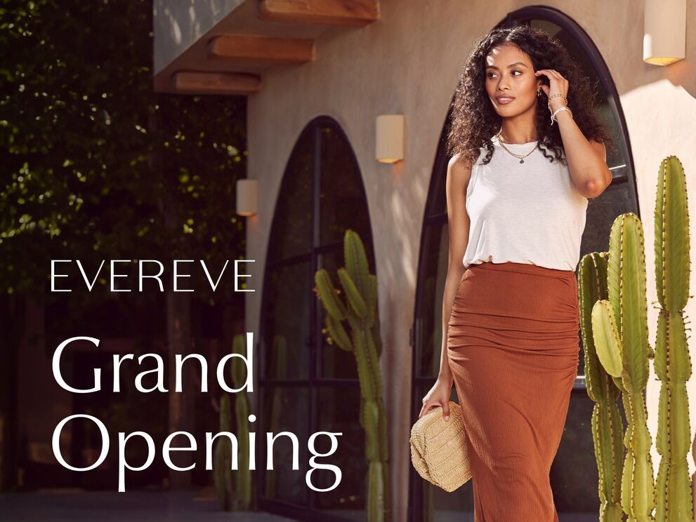 Brunette woman wearing a rust colored skirt and white blouse walking past a storefront with cactus plants promoting the opening of Evereve