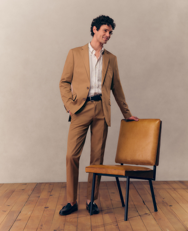 A man wearing a tan suit, wearing black dress shoes, leaning against a leather chair. 