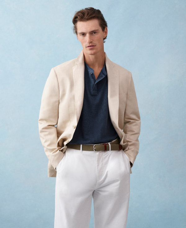 A man wearing white pants, with a blue shirt and a tan open jacket standing with his hands in his pocket.
