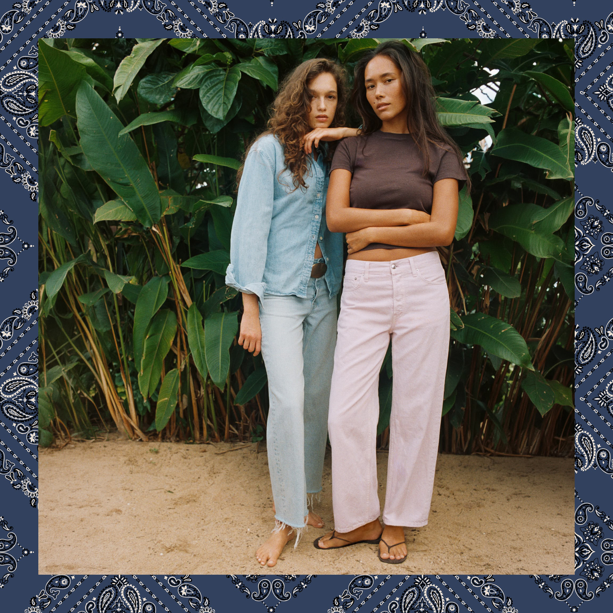 Two girls standing together in front of a greenery background. One girl is wearing jeans and a denim top and the other girl is wearing white jeans and a brown t shirt.